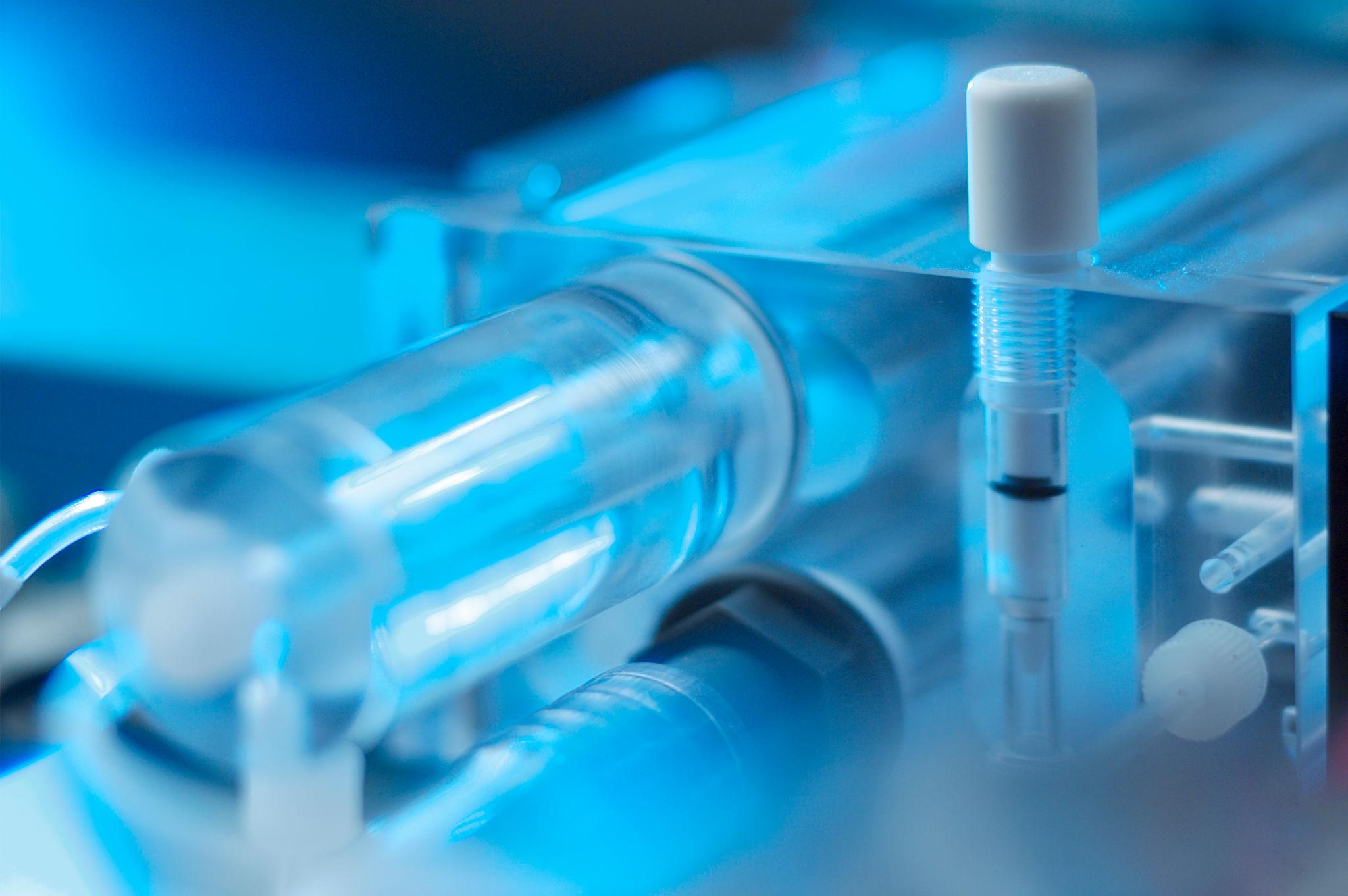 Close-up of a laboratory water treatment setup with clear tubes, a glass chamber, and a syringe-like component under blue lighting.